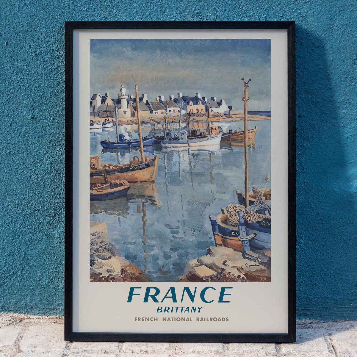 France Brittany Railway poster by Edmond Ceria featuring blue harbor boats, pale village roofs, cream sky, and coastal scenery, framed and displayed in home decor
